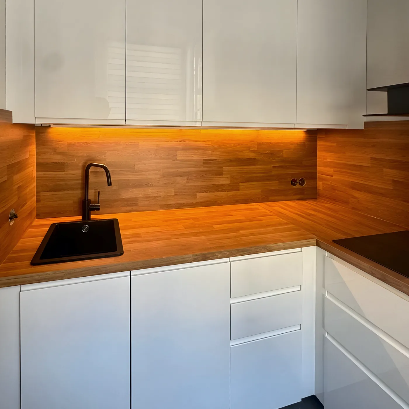 Weiße grifflose Küche mit beleuchteter Holzrückwand und schwarzer Spüle.